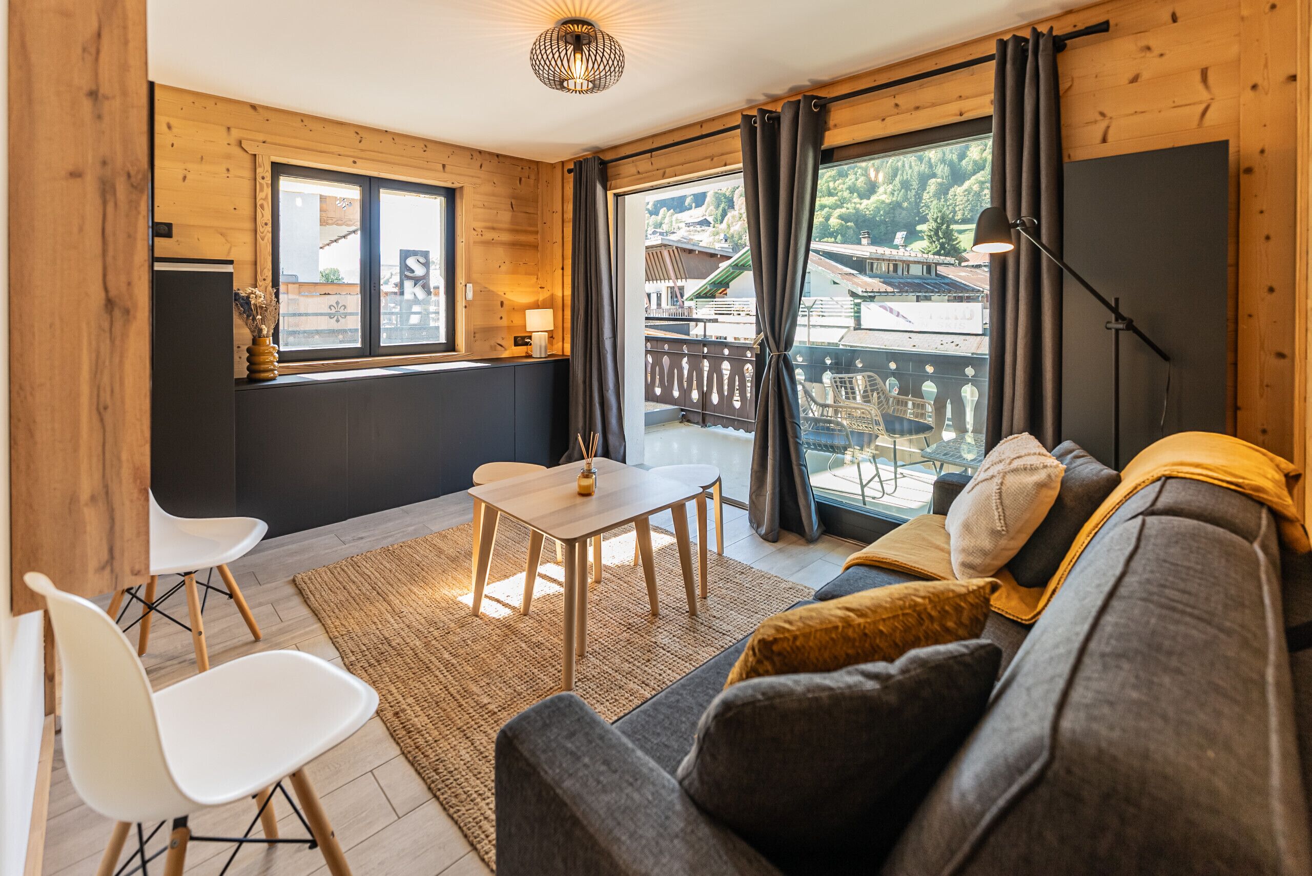 A cozy room with a sofa, a small table, and chairs. There is a window with curtains, a lamp, and a view of a balcony.