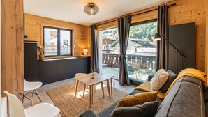 Ein gemütliches Zimmer mit einer Couch, einem kleinen Tisch und Stühlen. Ein Fenster mit Vorhängen, eine Lampe und ein Blick auf einen Balkon.