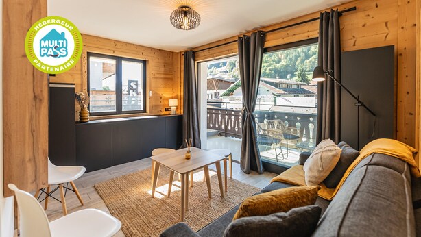 Ein gemütliches Zimmer mit einer Couch, einem kleinen Tisch und zwei Stühlen. Es gibt ein Fenster mit Vorhängen, eine Lampe und einen Blick auf einen Balkon.
