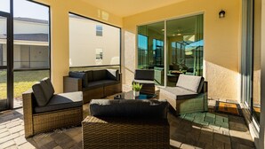 Un patio iluminado por el sol con muebles de mimbre, una puerta de cristal que da a un balcón y vista a un edificio al fondo.