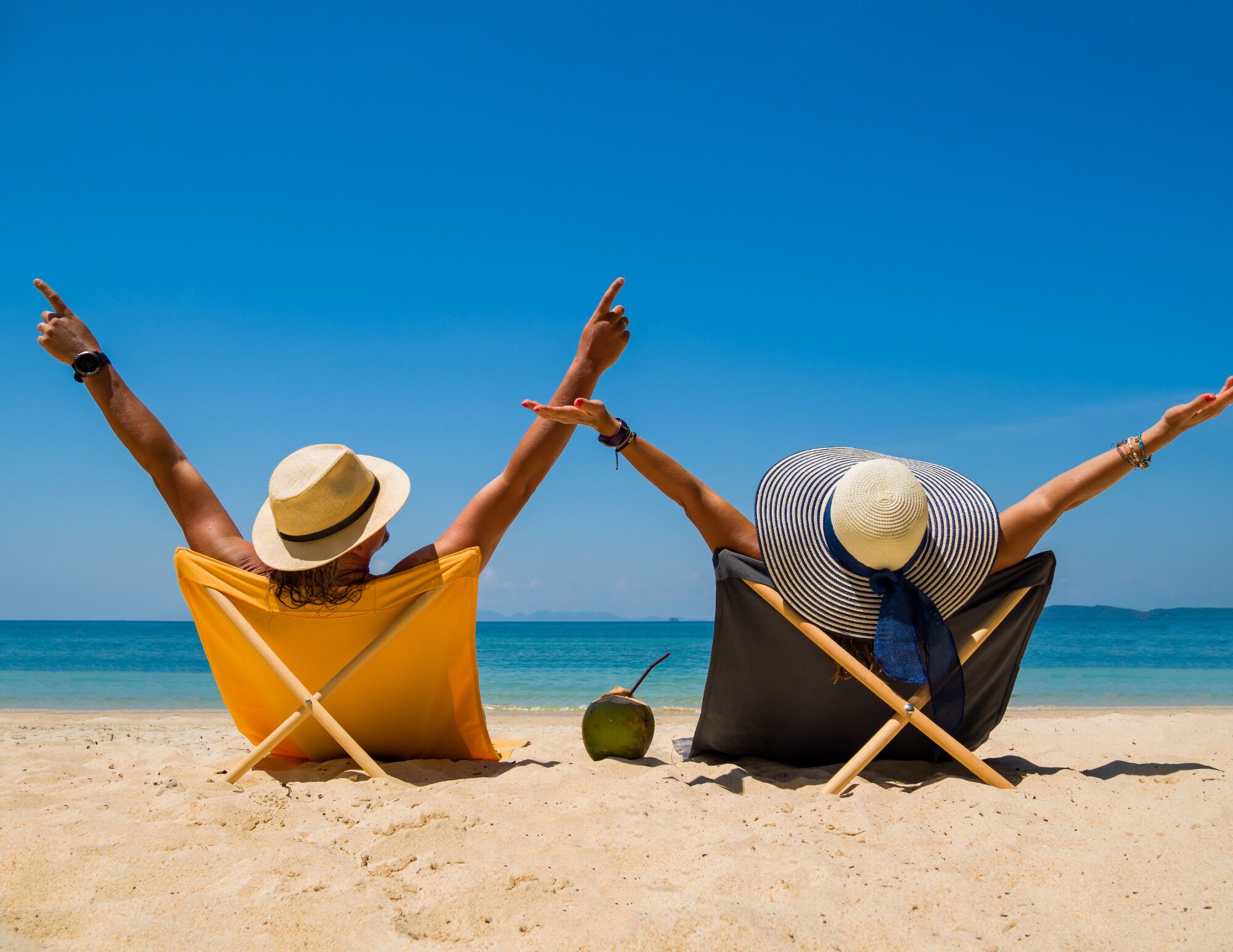 Two people sitting on beach chairs facing the sea, with their arms raised.