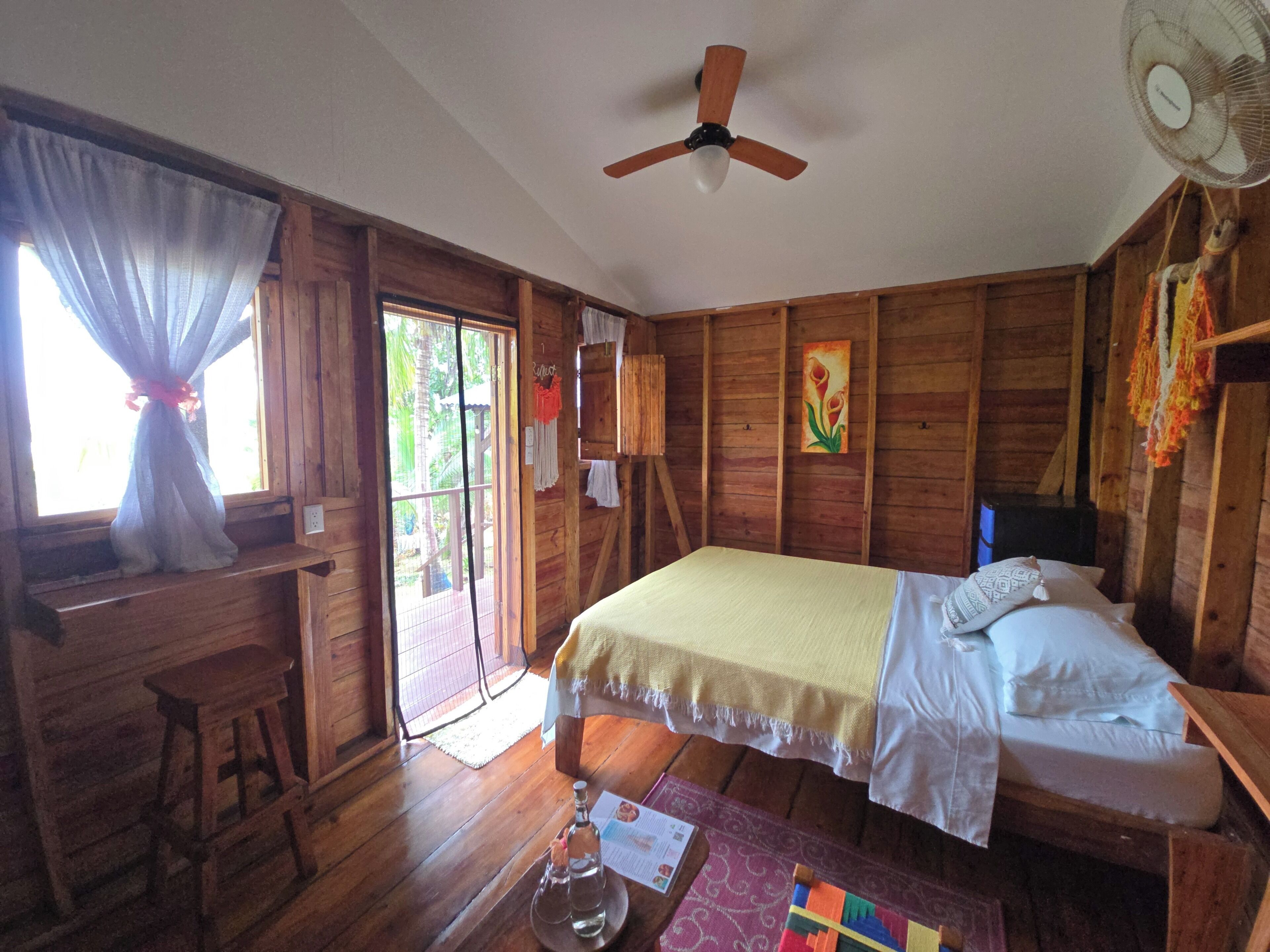 A wooden cabin room with a bed, a desk, a ceiling fan, and a window with curtains.