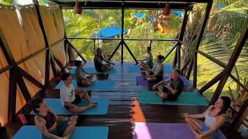 A group of people sitting on yoga mats in a covered outdoor area with a view of greenery.