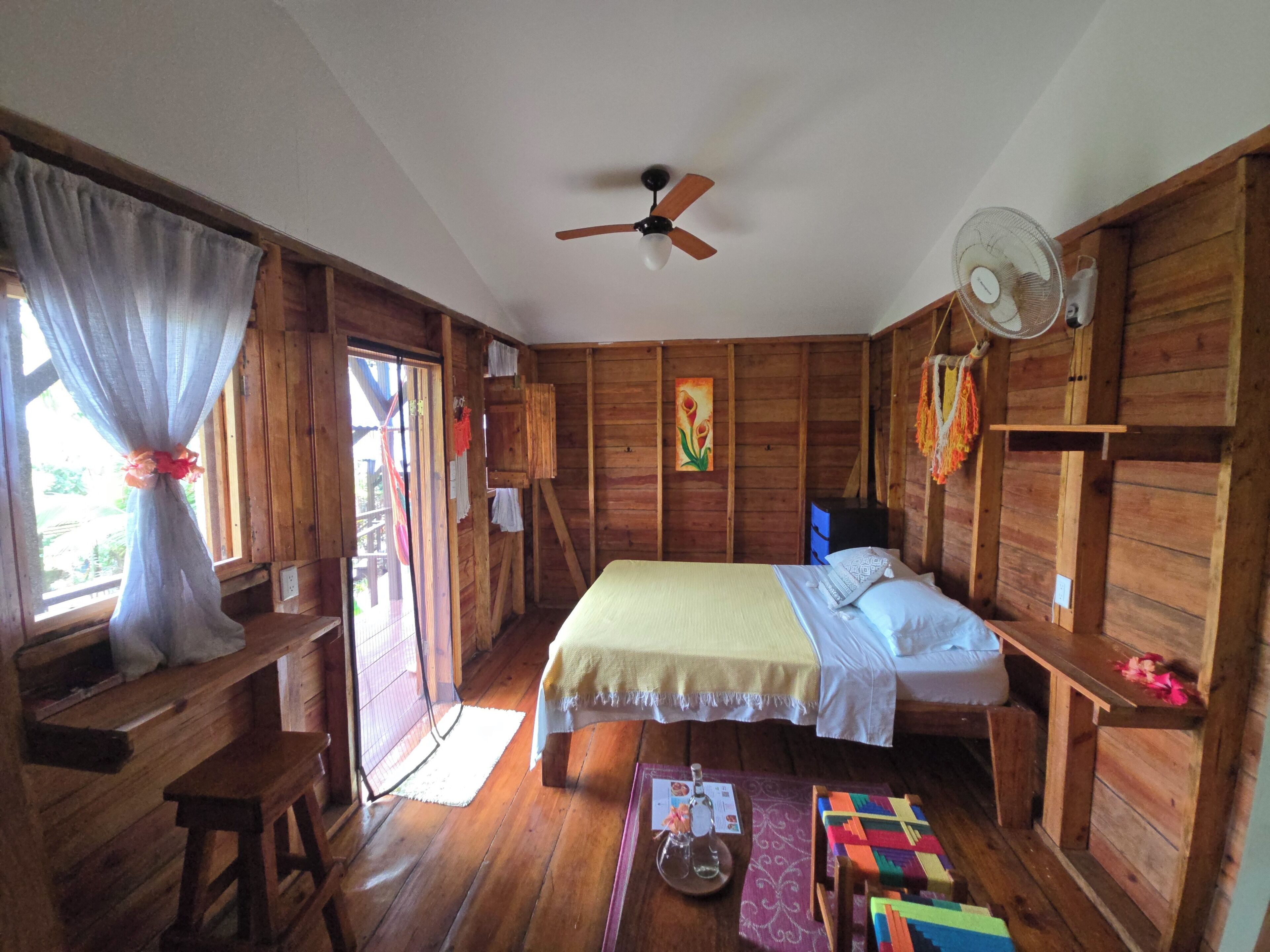 A wooden room with a bed, a ceiling fan, a window with curtains, and a wooden desk.