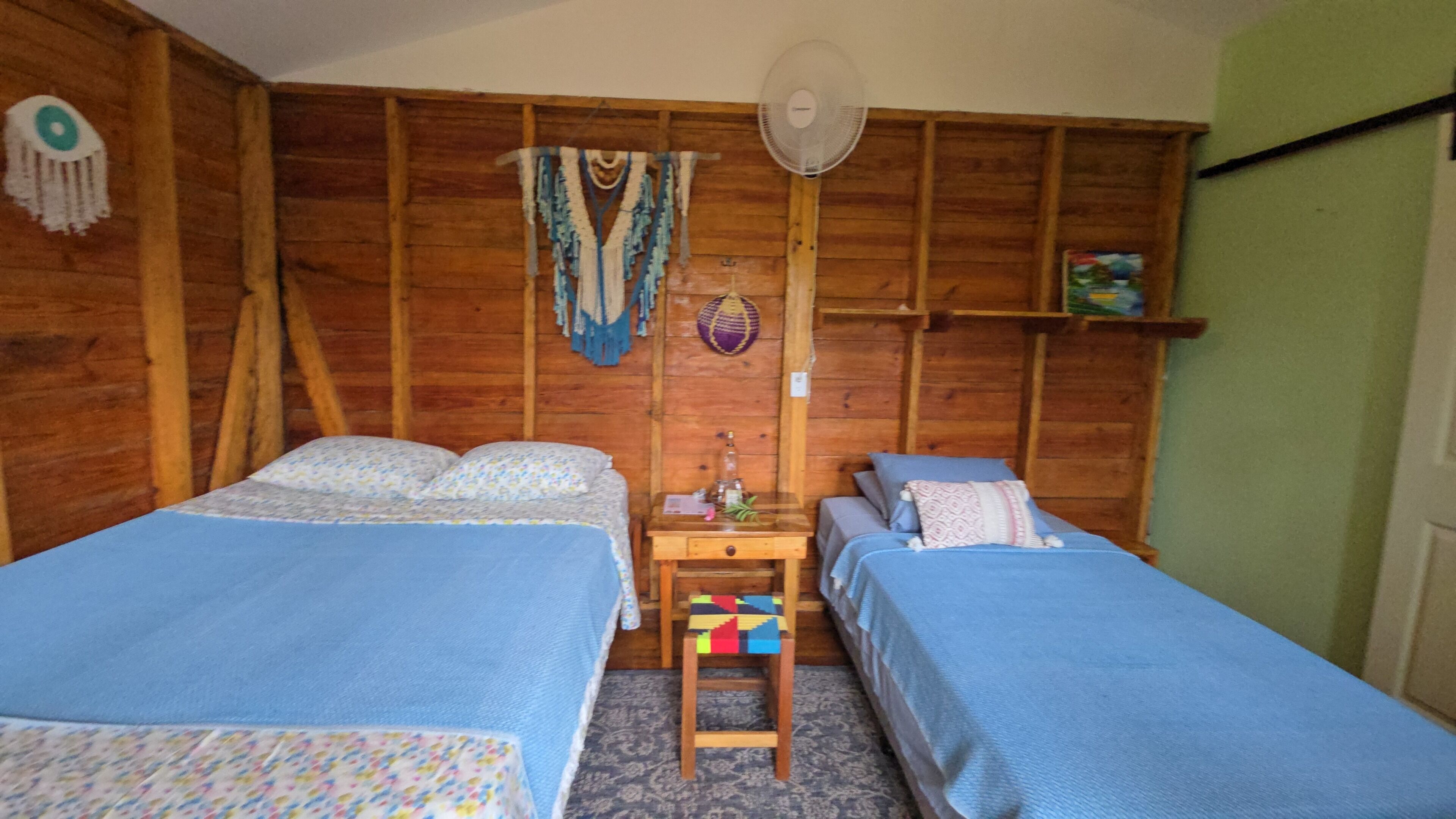 A room with two beds, a wooden wall, a fan, and a shelf with items.