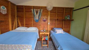 A room with two beds, a wooden wall, a fan, and a shelf with items.