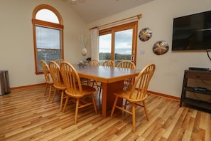 A dining area with a wooden table and chairs, large windows, and a TV on the wall.