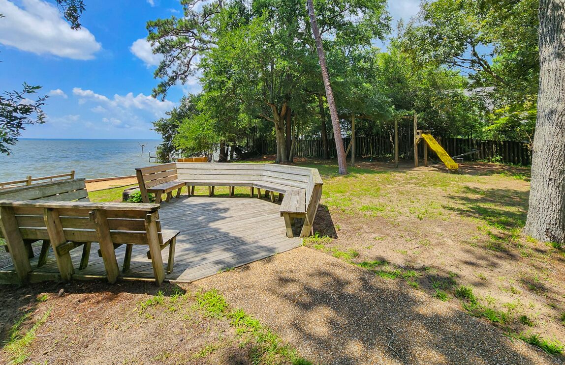 A wooden deck with benches overlooking a body of water, surrounded by trees and a playground.