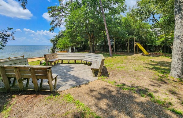 A wooden deck with benches overlooking a body of water, surrounded by trees and a playground.