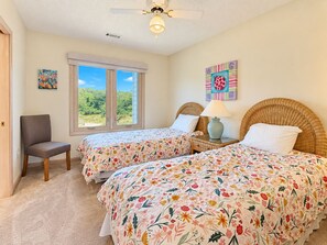 A bedroom with two beds, a chair, a ceiling fan, a window with a view, and artwork on the walls. - Carole's Castle - 4 BR Oceanside home in Southern Shores w/Private Pool (Southern Shores)