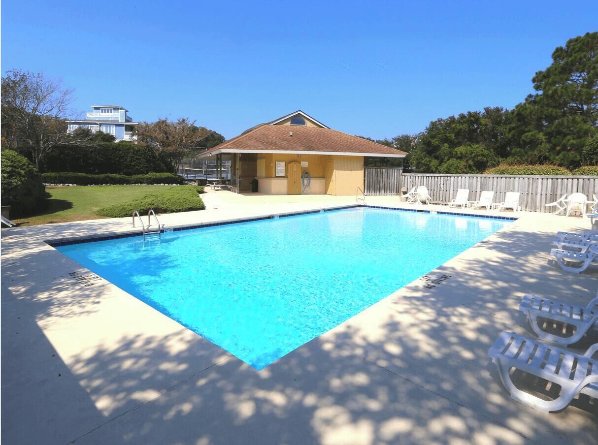 A community pool with a clear blue water surface, surrounded by a wooden deck and white lounge chairs.