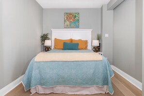 A bedroom with a bed, two bedside tables, a nightstand, a wall-mounted lamp, and a decorative wall piece.