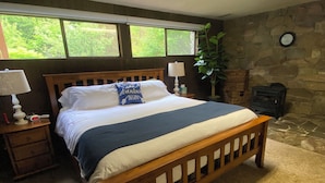A bedroom with a wooden bed, a nightstand with a lamp, a clock on the wall, and a stone wall. - Spacious Family Vacation Retreat! (Packwood)
