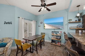 A dining area with a table and chairs, a ceiling fan, a TV displaying a beach scene, and a kitchen counter with a plant.