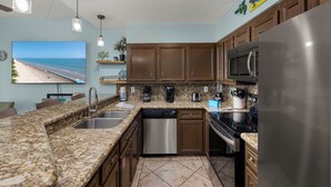 A kitchen with granite countertops, stainless steel appliances, wooden cabinets, and a beach-themed wall decal. - Beachside Bliss • Community Pool • 1 Min Walk to Beach (South Padre Island)