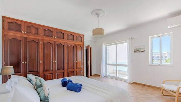 Un dormitorio con un armario grande, una cama con almohadas y una colcha, una silla y vistas al balcón exterior por una puerta corredera.