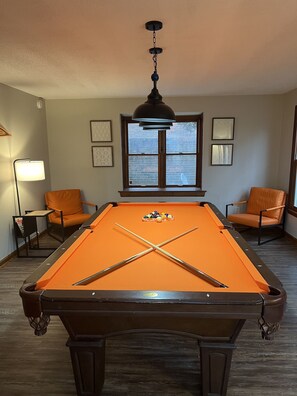 A pool table with balls, two chairs, a lamp, and a window with a view.