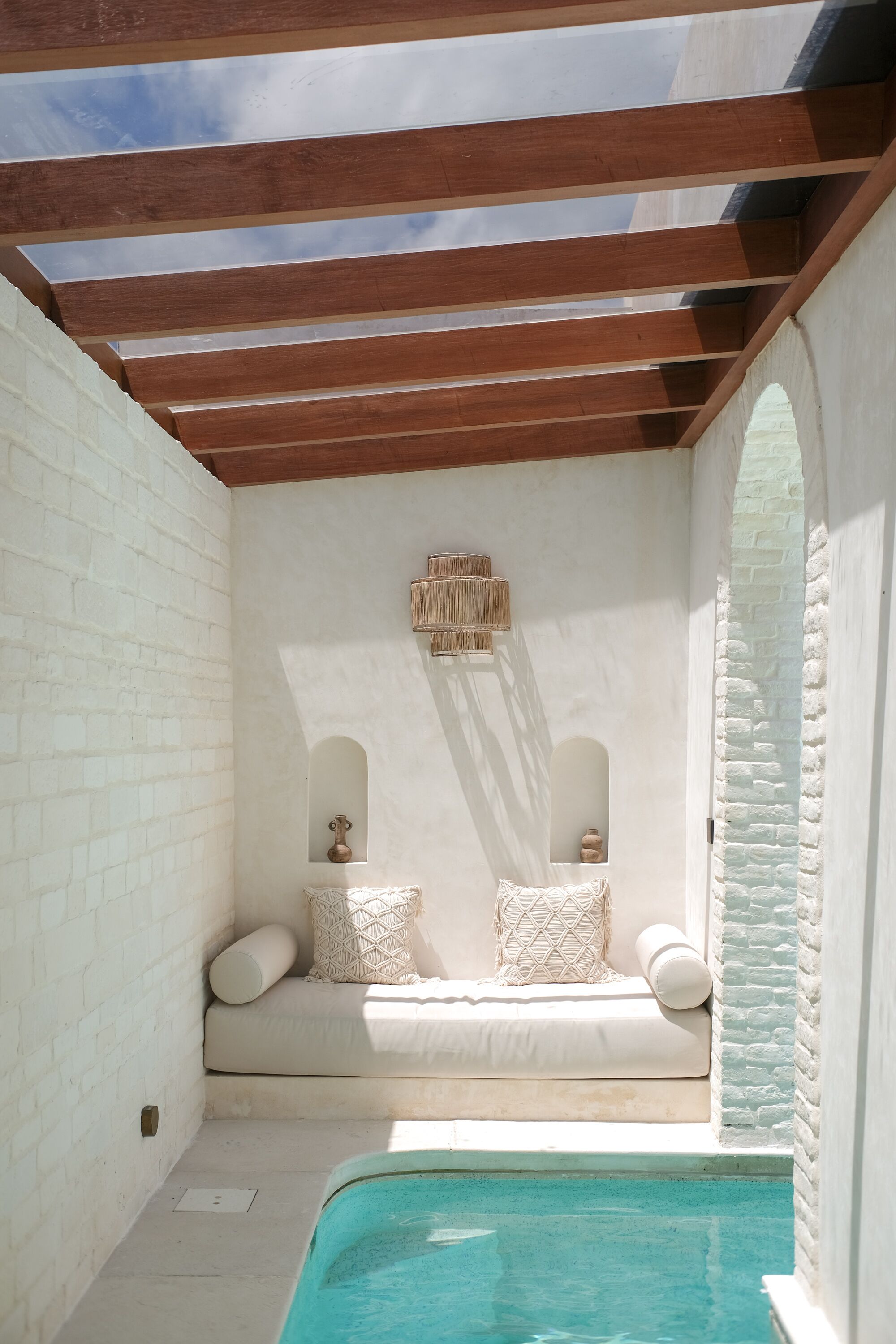 A spa area with a wooden ceiling, white brick walls, a small seating area with cushions, and a pool.