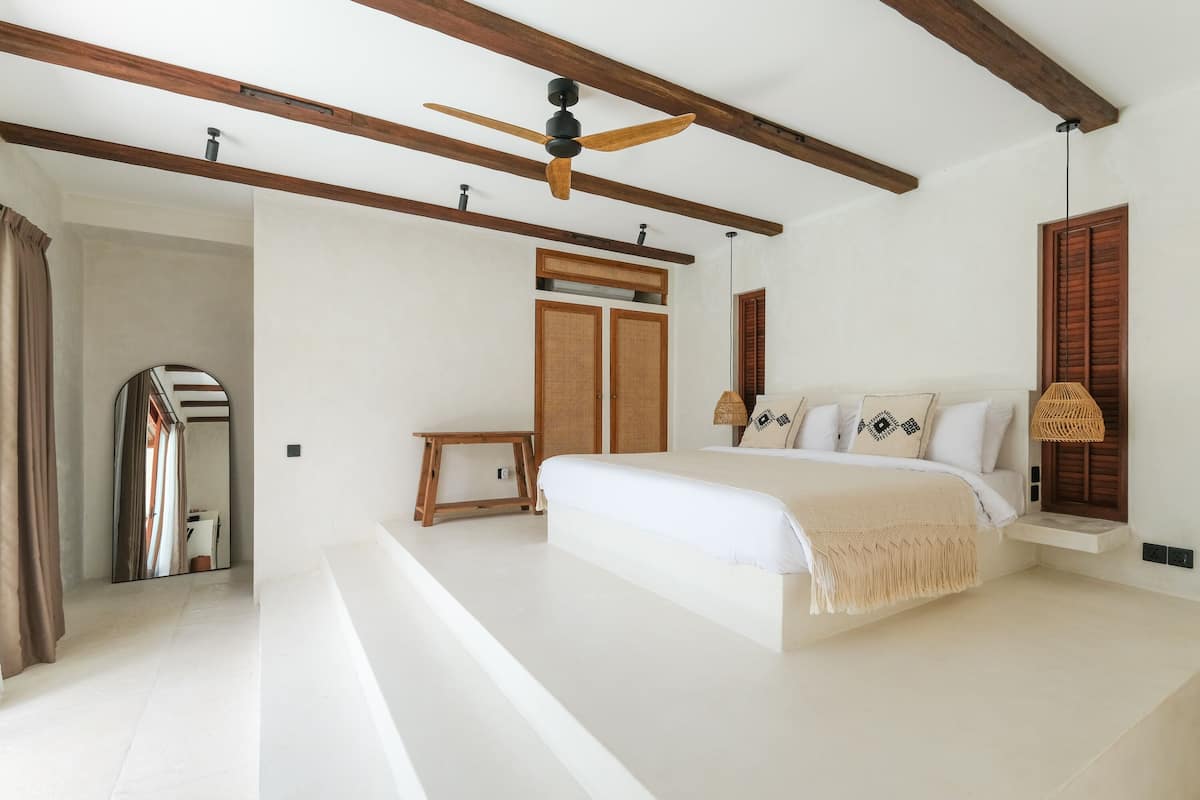 A spacious bedroom with a large bed, wooden beams, a ceiling fan, and a small bedside table.