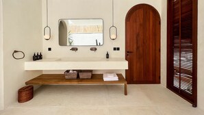 A modern bathroom with a white vanity, two pendant lights, a large mirror, and a wooden door. - Breathe Boutique (Pecatu)