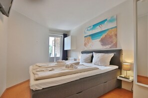 A hotel room with a large bed, a painting of a beach scene, a window, and a bedside table with a lamp.