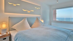 Un dormitorio con cama, almohadas, lámparas de noche, una mesita de noche, una obra de arte en la pared con pájaros volando y una ventana con vista al mar.