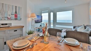 Comedor con mesa de madera dispuesta para dos personas, una botella de vino y un televisor al fondo.