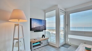 Una habitación de hotel moderna con un ventanal que ofrece vistas a la playa, un televisor de pantalla plana montado en la pared, una mesita de madera y una lámpara de pie.