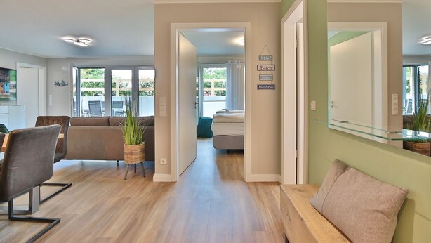 Un salón espacioso con sofá, una mesa de centro y una planta en maceta. Una puerta de cristal da acceso a un balcón con mobiliario exterior.