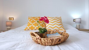 Un lit bien fait, avec une literie blanche, deux coussins décoratifs à motifs chevrons, un panier en osier contenant un livre et une plante en pot, et une lampe de chevet de chaque côté.