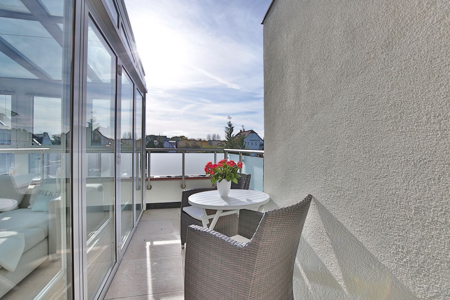 A balcony with a glass door, a white table, wicker chairs, and a potted plant with red flowers.