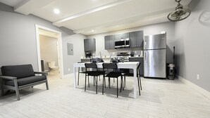 Une cuisine moderne avec des meubles gris, un réfrigérateur en acier inoxydable et un coin repas comprenant une table blanche et des chaises noires.