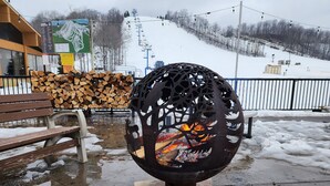 Une sculpture métallique dans laquelle brûle un feu, un banc en bois et une pile de bois de chauffage.