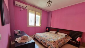 A bedroom with a bed, a desk with a lamp, and an air conditioner.