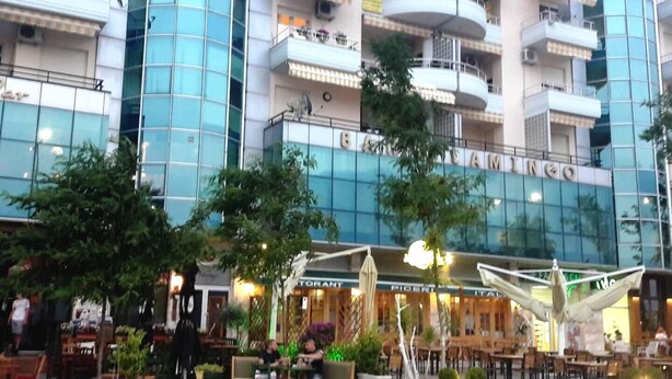 A multi-story hotel with a glass facade, outdoor seating area, and a restaurant named "PIGERIA ITALIA" at the ground level.