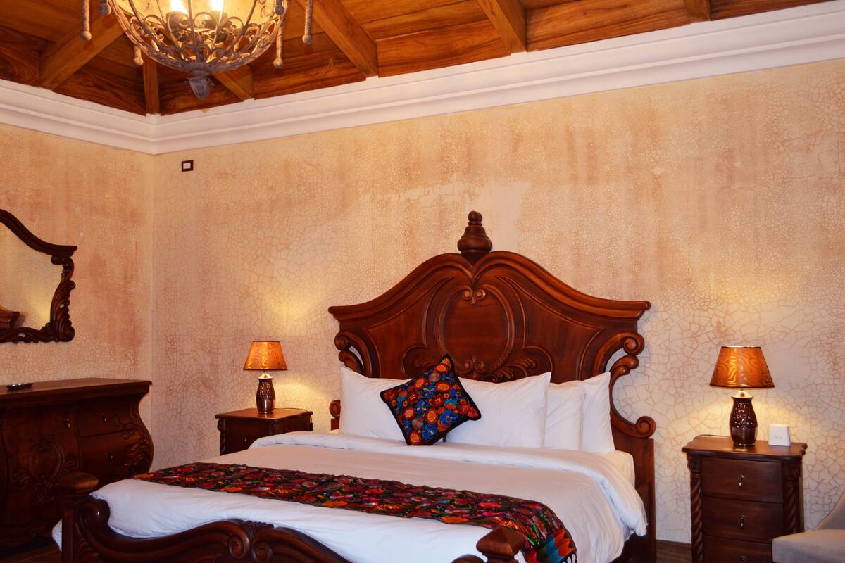 A bedroom with a wooden bed, two bedside tables with lamps, a nightstand, and a mirror on the wall.