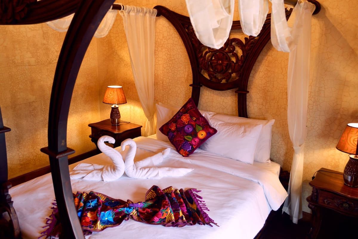 A four-poster bed with a canopy, white linens, a colorful pillow, and a decorative lamp on a wooden nightstand.