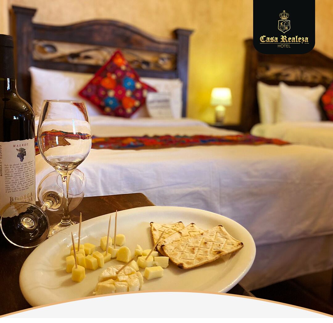 Habitación de hotel con cama, mesita de noche con botella de vino, copa de vino y plato con queso y galletas.