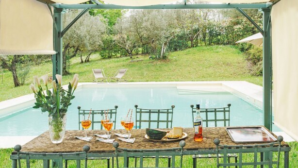 Ein Tisch mit Stühlen, eine Flasche, Gläser und eine Schüssel mit Essen, platziert unter einem Sonnensegel mit Blick auf einen Pool und Grünflächen.