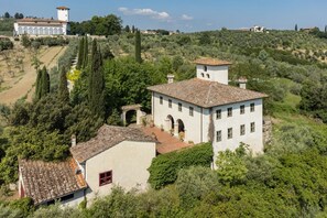 Un bâtiment blanc, doté d’un toit en tuiles, entouré de verdure et d’oliviers, avec une structure en forme de tour en arrière-plan.