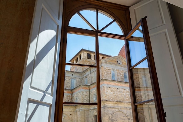 A room with a large arched window, a wooden door frame, and a view of a historic building with multiple windows and a red roof.