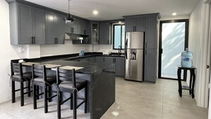 Een moderne keuken met grijze kastjes, een centraal eiland met barkrukken, een roestvrijstalen koelkast en een waterdispenser.