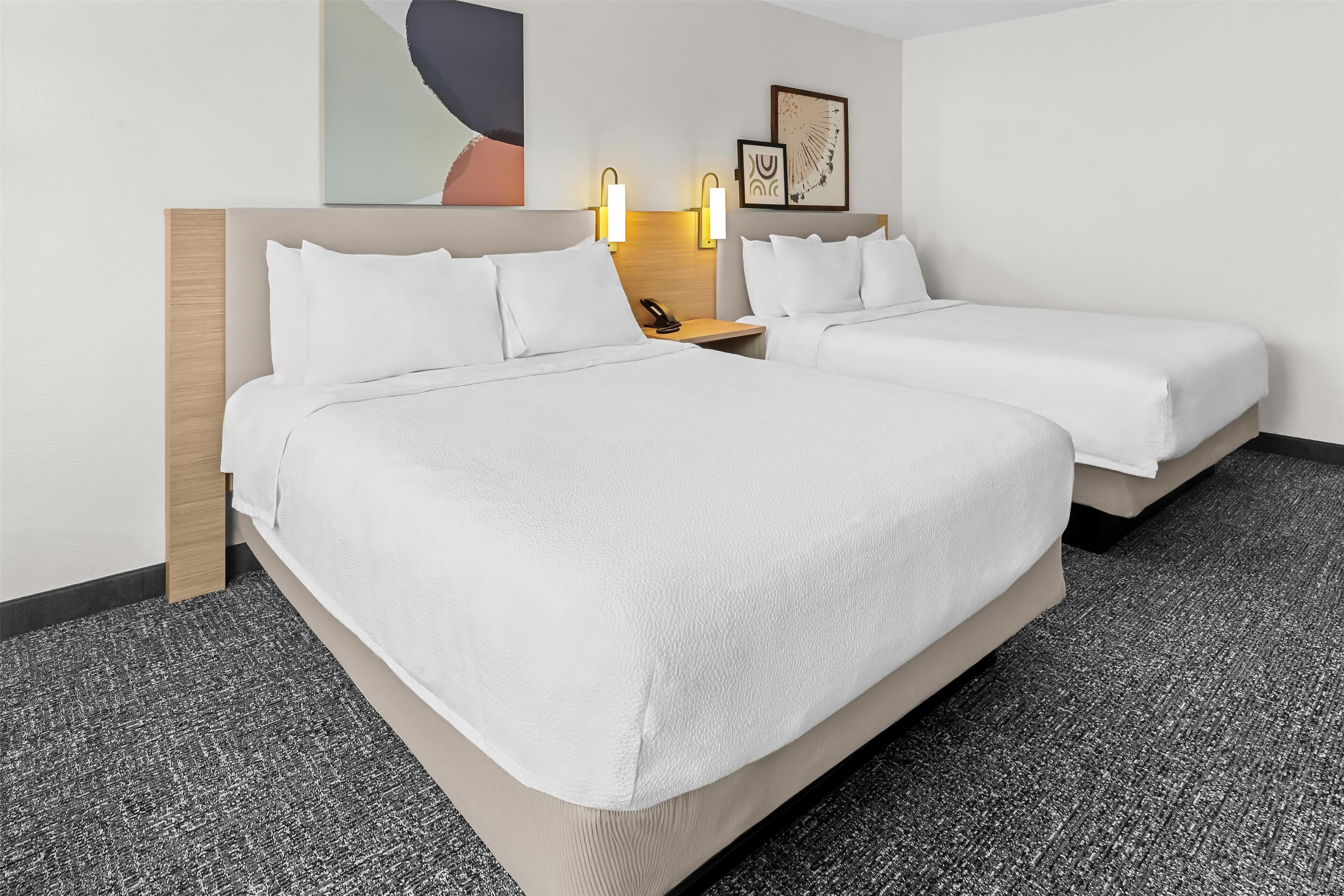 Quarto de hotel com duas camas, uma cabeceira de madeira, uma mesinha de cabeceira com telefone e luminárias fixadas na parede.