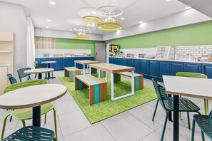 A modern kitchen with blue cabinets, green and white countertops, and a green accent wall. There are multiple tables and chairs, and a ceiling with circular light fixtures. - Spark by Hilton Sumter (Sumter)