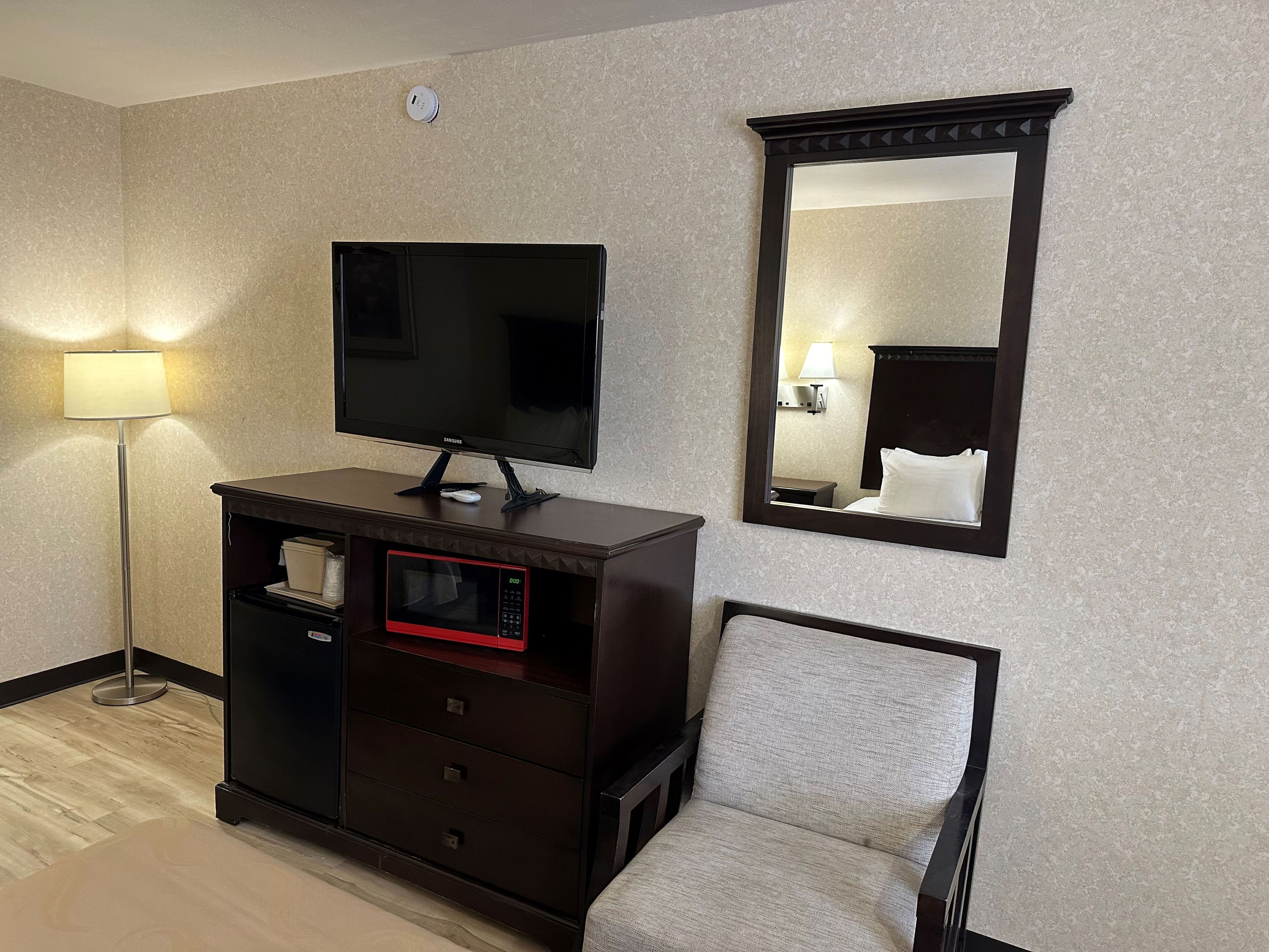 Ein Hotelzimmer mit Flachbildfernseher auf einer Holzkommode, einer Mikrowelle, einem Sessel und einem großen Spiegel an der Wand.