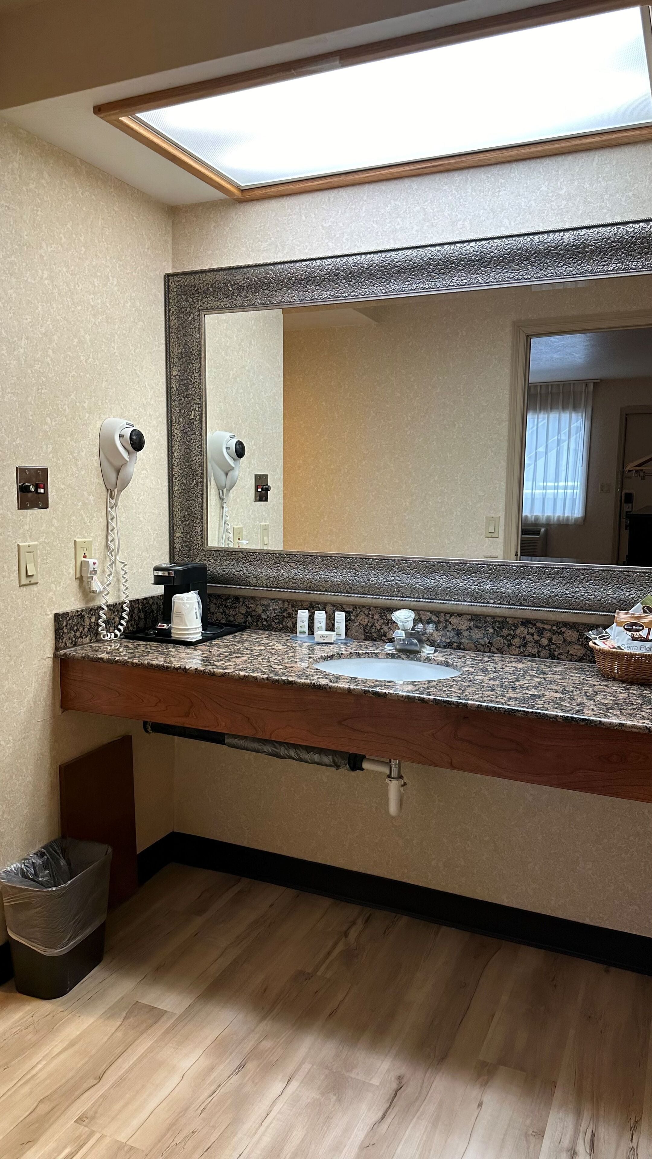 A hotel bathroom with a granite countertop, a sink, a mirror, a hairdryer, a coffee maker, and a trash bin.