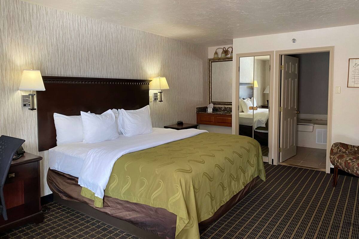 Una habitación de hotel con una cama grande, dos lámparas de noche, una mesita de noche, un escritorio, una silla, un espejo y una puerta que da a un baño.