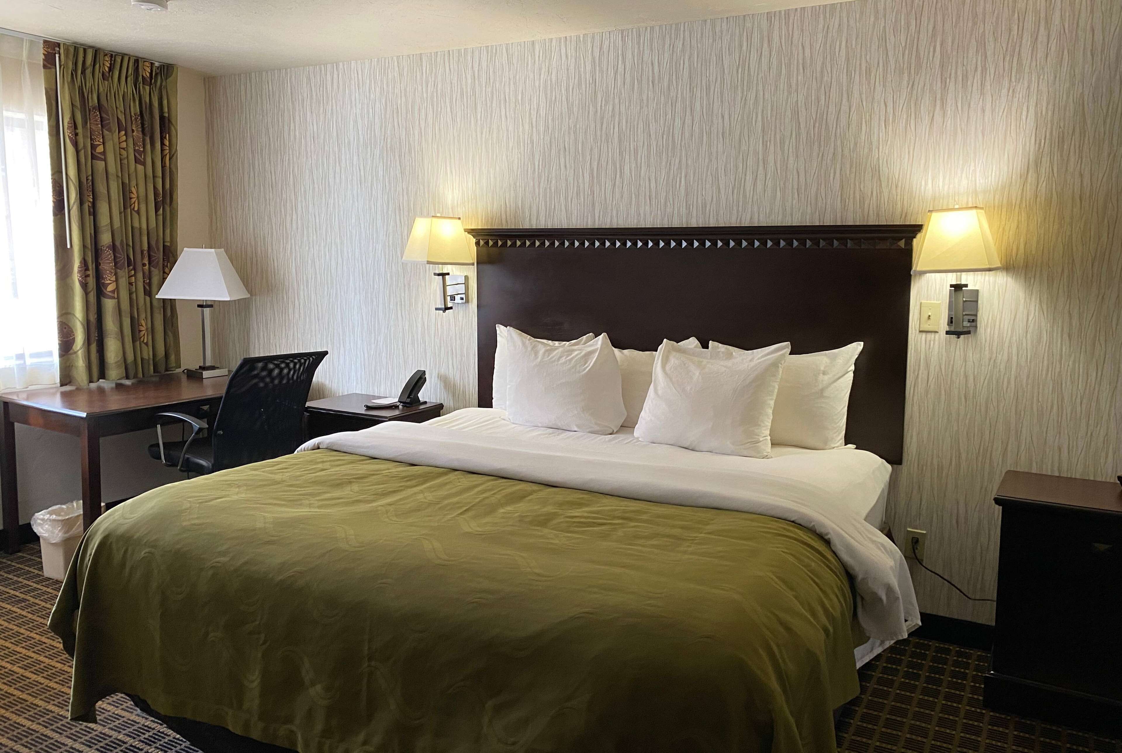 A hotel room with a large bed, two bedside lamps, a desk with a chair, and a telephone.