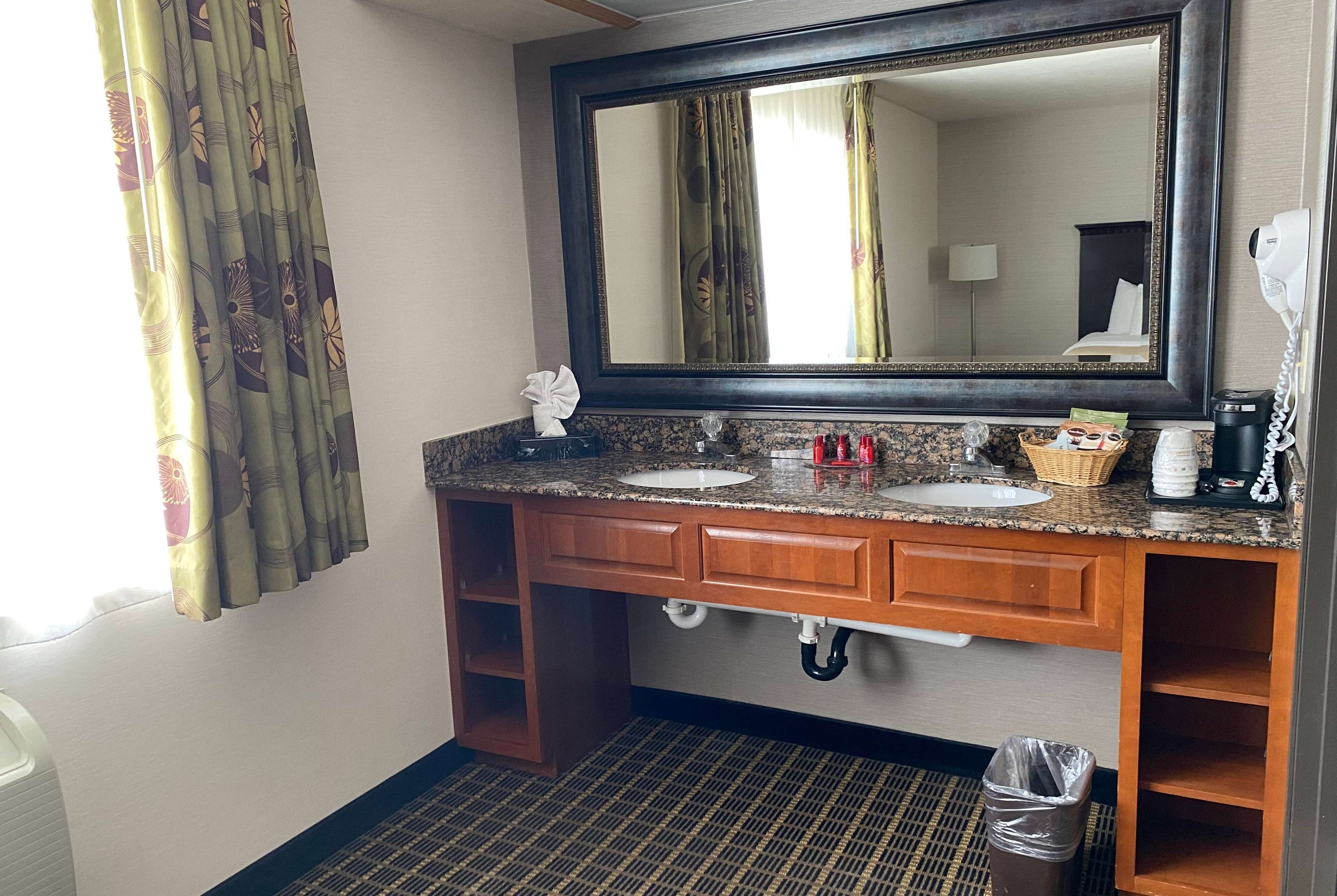 Ein Badezimmer in einem Hotel mit zwei Waschbecken, einem großen Spiegel, einem Handtuchspender, einem Mülleimer und einem an der Wand befestigten Föhn.