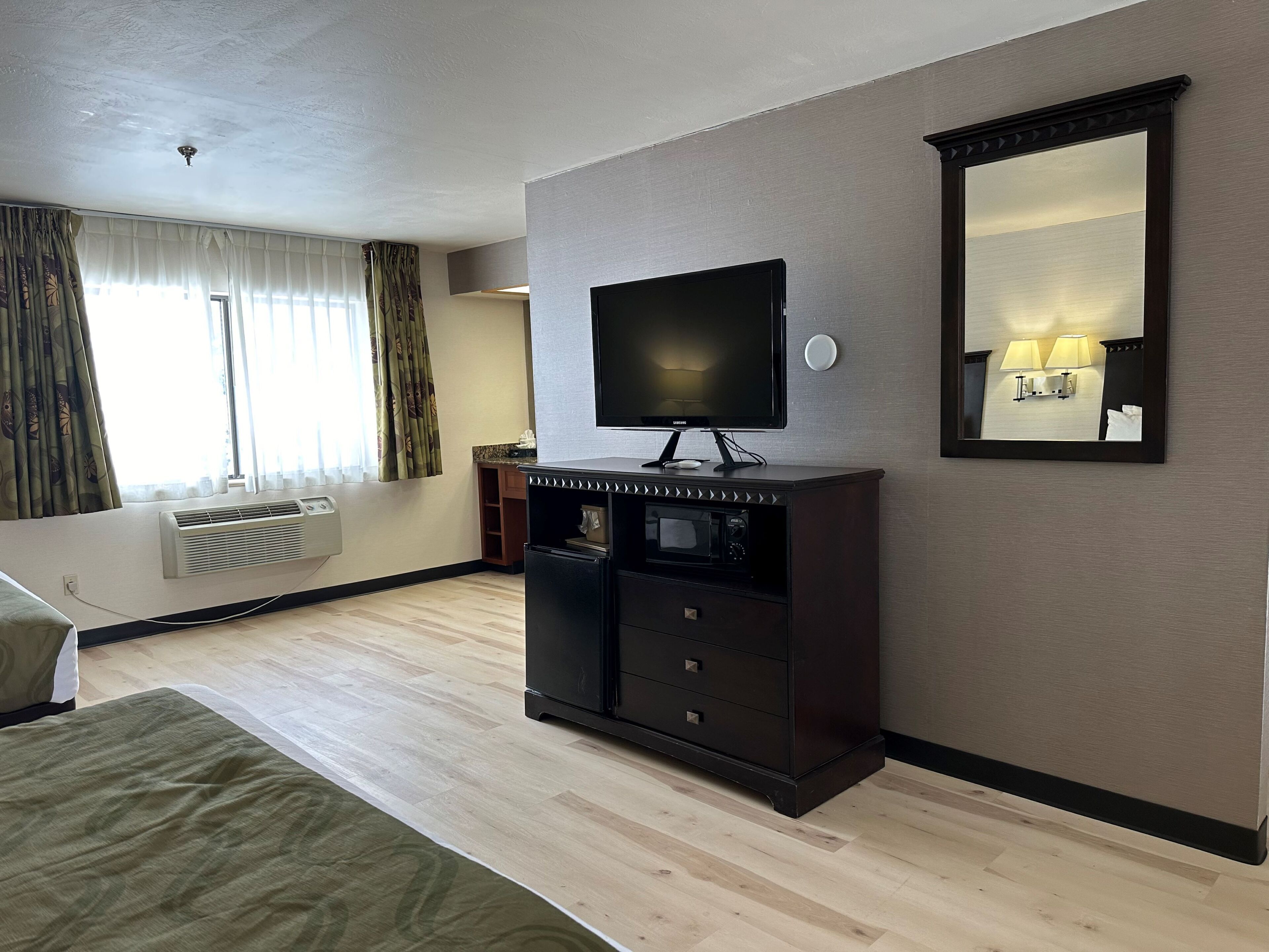 A hotel room with a flat-screen TV on a wooden cabinet, a bed with a green bedspread, an air conditioning unit, and a large window with curtains.